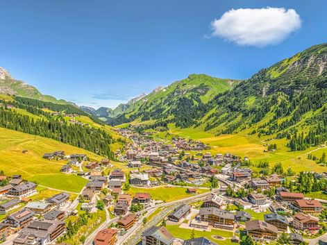 Luftaufnahme des Dorfes Lech am Arlberg in einem grünen Tal, umgeben von bewaldeten Bergen unter blauem Himmel mit vereinzelten Wolken.