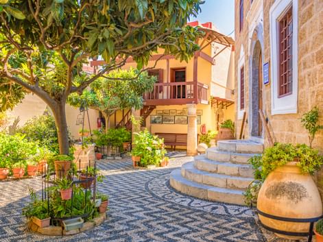 Sonniger Innenhof in der Altstadt von Rhodos mit gemustertem Steinpflaster, geschwungenen Stufen, Topfpflanzen, einem Baum und traditionellen Gebäuden.