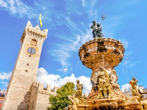 Neptunbrunnen in Trient mit kunstvollen Skulpturen und Wasserfontänen. Ein historischer Uhrturm mit Zinnen steht im Hintergrund unter blauem Himmel.