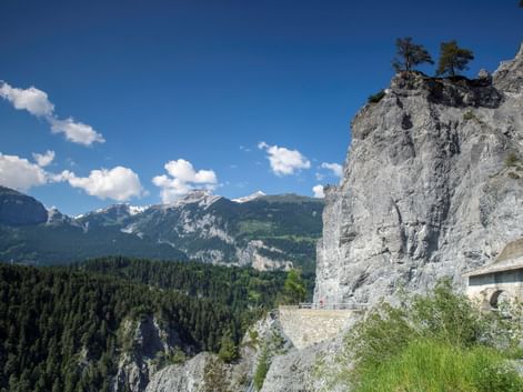 Rheinschlucht Ruinaulta mit steilen grauen Felswänden, Bergstraße und bewaldeten Bergen unter blauem Himmel. Schneebedeckte Gipfel in der Ferne.