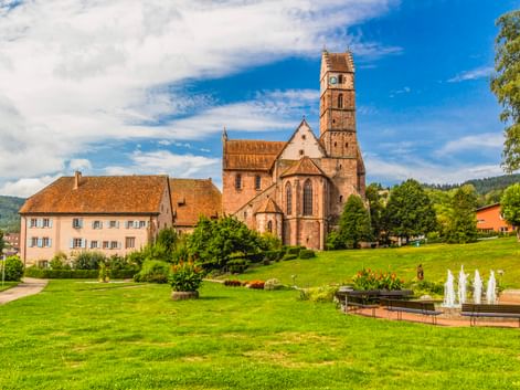 Kloster Alpirsbach mit rotem Sandsteinkirchturm und angrenzenden Gebäuden auf grüner Wiese mit Brunnen, umgeben von Bäumen und Hügeln.