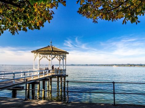 Holzsteg mit weißem Pavillon am Bodensee bei Bregenz. Menschen sitzen unter der überdachten Struktur, mit ruhigem blauen Wasser und ferner Küstenlinie.