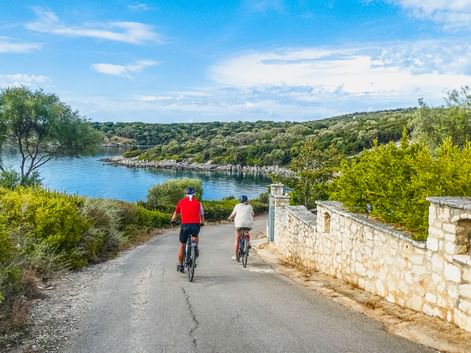 Zwei Radfahrer auf einer asphaltierten Straße neben einer Steinmauer mit blauer Bucht und grünen Hügeln der Insel Meganisi im Hintergrund.