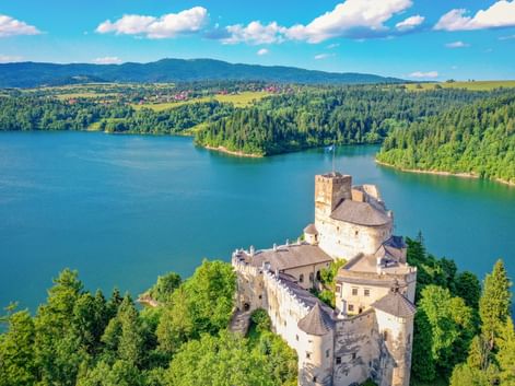 Luftaufnahme der Burg Niedzica auf einer bewaldeten Halbinsel, umgeben von türkisfarbenem Seewasser, mit Bergen und Wäldern im Hintergrund.