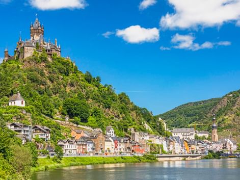 Stadt Cochem an der Mosel mit der Reichsburg auf einem grünen Hügel. Bunte Häuser säumen das Flussufer unter blauem Himmel.