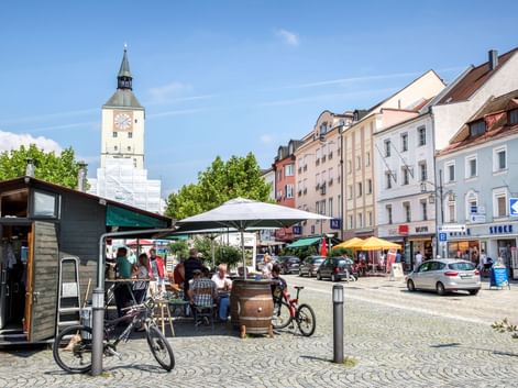 Kopfsteinpflaster-Marktplatz in Deggendorf mit weißem Uhrturm, bunten Gebäuden, Marktständen, geparkten Fahrrädern und Außensitzplätzen unter blauem Himmel.