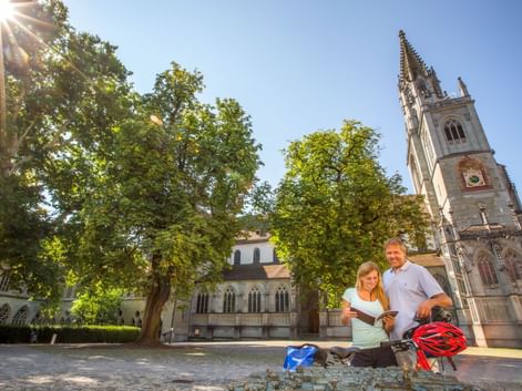 Zwei Radfahrer mit Rädern und Helmen ruhen an einem Brunnen vor dem Konstanzer Münster. Der gotische Kirchturm erhebt sich hinter grünen Bäumen unter blauem Himmel.