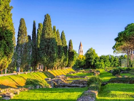 Archäologische Stätte in Aquileia mit antiken Steinruinen auf grünen Rasenflächen, hohen Zypressen und dem Glockenturm der Basilika im Hintergrund.