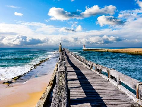 Holzsteg mit Geländer führt ins Meer in Capbreton, mit einem Leuchtturm am Ende und einer weißen Bake auf der rechten Mole.