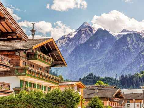 Traditionelle Alpenchalets in Kaprun mit blumengeschmückten Balkonen vor schneebedeckten Berggipfeln unter blauem Himmel mit weißen Wolken.