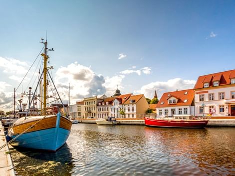 Harbor of Ueckermünde with blue wooden fishing boat in foreground and red boat behind. Historic buildings with orange-tiled roofs line the waterfront.