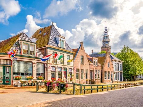 Traditionelle niederländische Gebäude mit bunten Flaggen entlang eines Kanals in Bolsward. Ein Kirchturm erhebt sich im Hintergrund unter blauem Himmel.