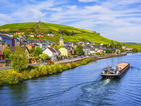 Frachtschiff auf der Mosel vor Wormeldange mit bunten Häusern, Kirchturm und weinbewachsenen Hügeln unter blauem Himmel.