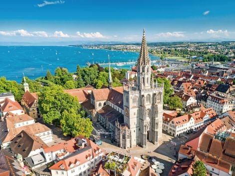 Luftaufnahme des Konstanzer Münsters mit gotischem Turm über den rotgedeckten Gebäuden. Bodensee mit Segelbooten im Hintergrund sichtbar.