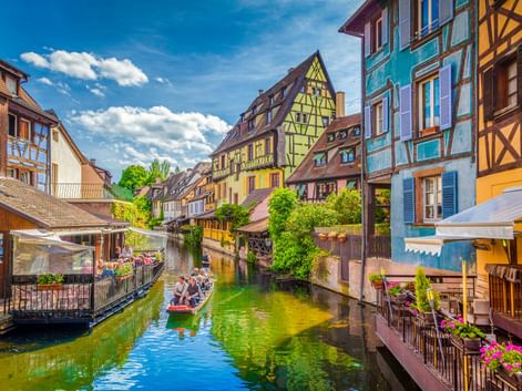 Bunte Fachwerkhäuser säumen einen Kanal in Klein-Venedig, Colmar. Ein Boot mit Touristen schwimmt auf dem grünen Wasser unter blauem Himmel.