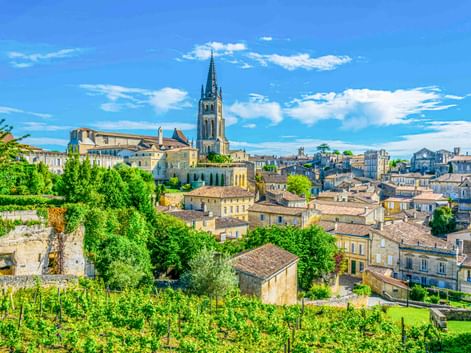 Panoramablick auf Saint-Émilion mit Steingebäuden, einem markanten Kirchturm und üppiger grüner Vegetation unter blauem Himmel mit weißen Wolken.