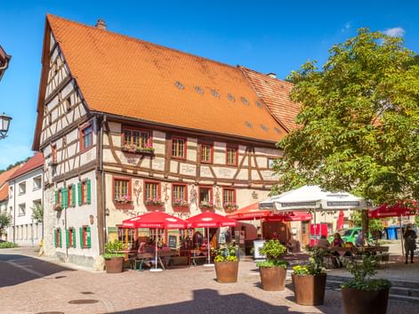Traditional half-timbered building with orange tile roof and outdoor café with red umbrellas in Fridingen old town square under blue sky.