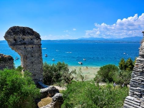 Antike Steinturmruinen in Sirmione mit türkisfarbenem Gardasee, Booten, Strand und Bergen unter blauem Himmel.