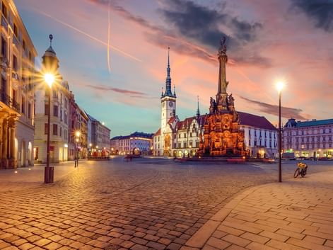 Olmützer Marktplatz in der Dämmerung mit beleuchteter Dreifaltigkeitssäule und Rathausturm. Kopfsteinpflaster und historische Gebäude unter buntem Himmel.