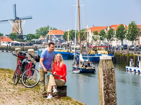 Paar mit Fahrrädern am Hafen in Zierikzee. Traditionelle Windmühle, Boote und historische Gebäude mit orangen Dächern im Hintergrund.