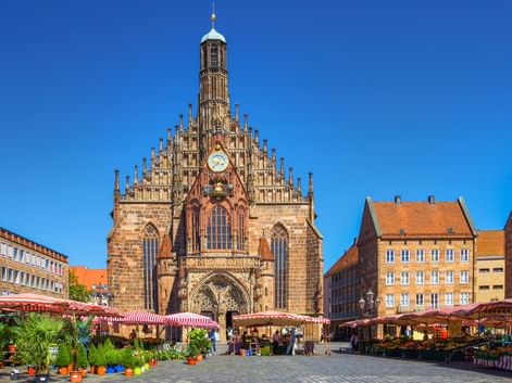 Frauenkirche church at Nuremberg's Hauptmarkt square with market stalls. Gothic facade with ornate gables, clock, and tower against blue sky.