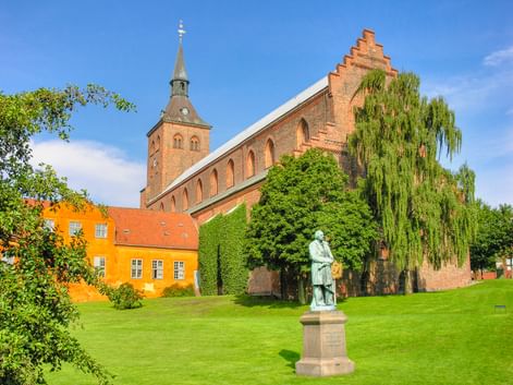 Bronzestatue von H.C. Andersen auf grüner Wiese in Odense, mit rotem Backsteinkirchturm und gelbem historischen Gebäude im Hintergrund.