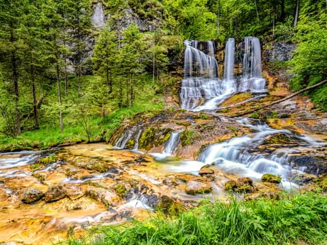 Wasserfälle Weissbach in Schneizlreuth, die über orange-braune Felsen inmitten von üppigem grünem Wald und moosbewachsenen Steinen fließen.