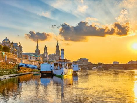 Sunset view of Dresden's Brühlsche Terrasse with boats moored on the Elbe River. Historic buildings and church towers visible against golden sky.