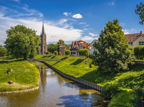 Kanal mit grünen Böschungen im alten Hafen von Rinteln. Kirchturm der St. Sturmius Kirche und Fachwerkhäuser zwischen Bäumen unter blauem Himmel.