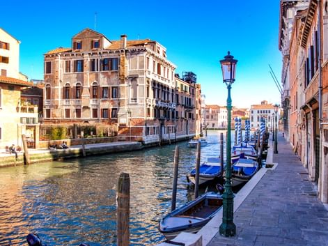 Venezianischer Kanal mit vertäuten Gondeln entlang eines Gehwegs. Historische Gebäude säumen den Wasserweg unter blauem Himmel. Traditionelle Laternen stehen entlang der Promenade.