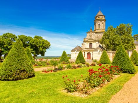 Abteikirche mit achteckigem Turm in Saint-Florent-le-Vieil, umgeben von formalen Gärten mit kegelförmigen Hecken, bunten Blumenbeeten und Bäumen.