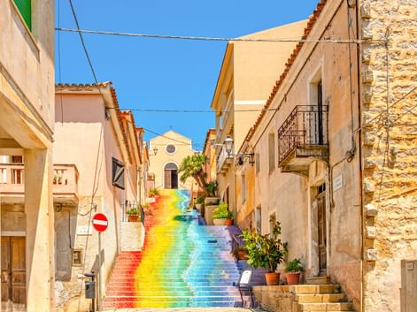 Bunte Regenbogentreppe in einer engen Gasse in Arzachena, Sardinien, gesäumt von traditionellen beigen Gebäuden unter blauem Himmel.