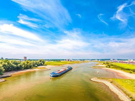 Luftaufnahme eines blauen Frachtschiffs auf dem Niederrhein bei Arnheim. Grüne Flussufer und städtische Gebäude unter blauem Himmel.