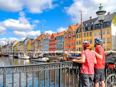 Zwei Radfahrer mit Helmen betrachten bunte historische Gebäude entlang des Nyhavn-Hafens in Kopenhagen. Traditionelle Boote liegen im Kanal.