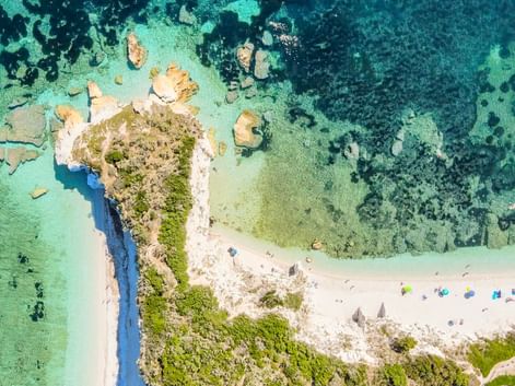 Luftaufnahme der Badebucht Padulella auf Elba mit weißem Sand, türkisfarbenem Wasser, felsiger Küste, grüner Vegetation und bunten Sonnenschirmen.