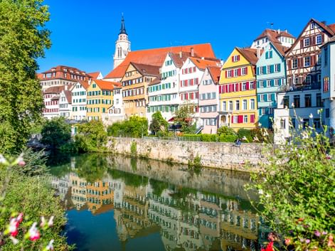 Row of colorful half-timbered houses along the Neckar River in Tübingen, with a church tower and reflections in the water, framed by green foliage.