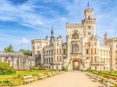 Weißes Schloss Hluboka mit zinnenbesetzten Türmen und gotischer Architektur. Ein Kiesweg führt durch formale Gärten mit Hecken.