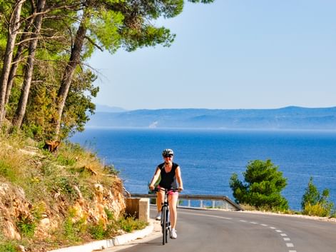 Radfahrerin mit Helm auf Küstenstraße in Kroatien. Blaues Meer und ferne Berge sichtbar, Kiefern säumen den Hang.