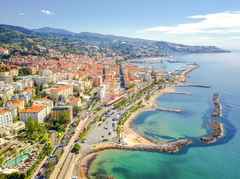 Panoramablick auf Sanremo, Italien, mit Küstenstadt, Stränden, Hafen, palmengesäumter Promenade und Bergen im Hintergrund.