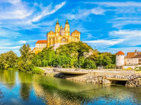 Yellow baroque Melk Abbey with twin towers sits atop a green hill above the Danube River. A curved promenade lines the riverbank under blue sky.