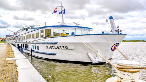 Weißes Flusskreuzfahrtschiff MS Rigoletto mit blauem Rand am Betonpier vertäut. Das Schiff hat große Fenster und Flaggen auf dem Oberdeck.