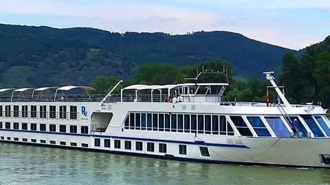 Weißes Flusskreuzfahrtschiff MS SE-Manon mit blauem Streifendesign fährt auf ruhigem Wasser vor grünen Bergen.