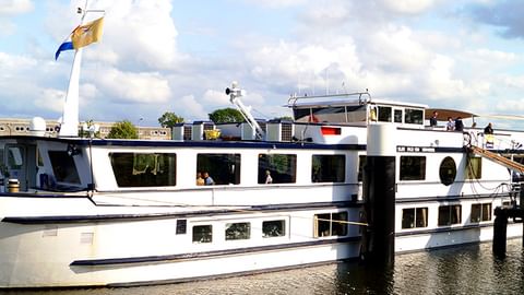 Weißes Flusskreuzfahrtschiff MS Olympia am Kai mit wehenden Flaggen. Weitere Schiffe im Hintergrund unter bewölktem Himmel.