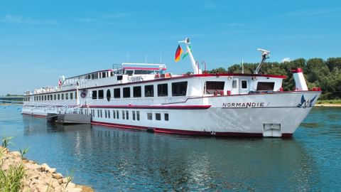 Weißes Flusskreuzfahrtschiff MS Normandie mit rotem Rand fährt auf blauem Wasser unter klarem Himmel, grüne Bäume und felsiges Ufer sichtbar.