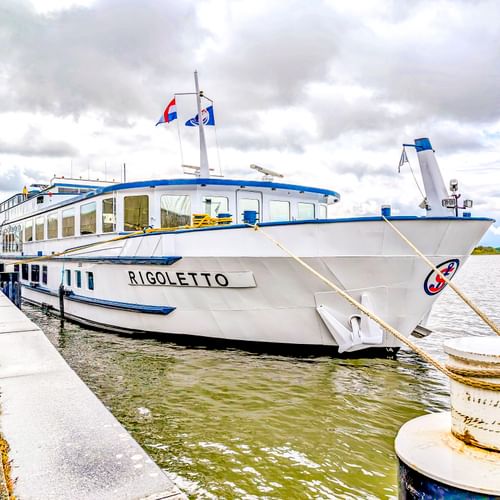 Weißes Flusskreuzfahrtschiff MS Rigoletto mit blauem Rand am Betonpier vertäut. Das Schiff hat große Fenster und Flaggen auf dem Oberdeck.