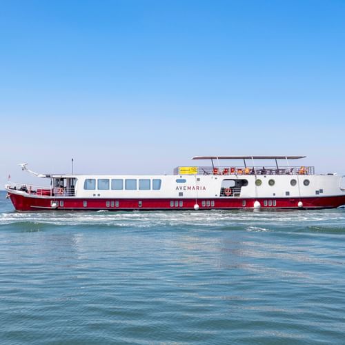 MS Ave Maria river cruise ship with red and white hull sailing on calm blue water under clear sky. The vessel features multiple decks and portholes.