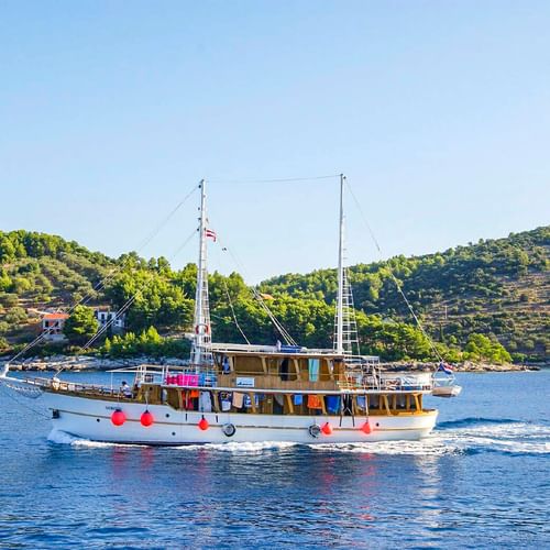 Motorschiff MS Dalmatino mit roten Fendern auf ruhigem blauen Wasser. Grüne terrassierte Hügel mit vereinzelten Gebäuden im Hintergrund.