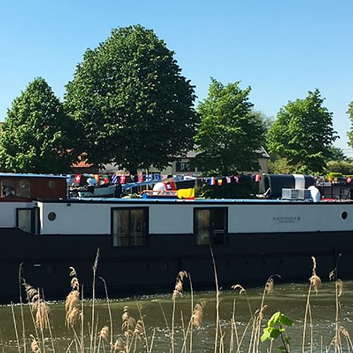 MS Magnifique III, a dark-hulled river cruise ship, moored along a green riverbank with trees and buildings in the background under blue sky.