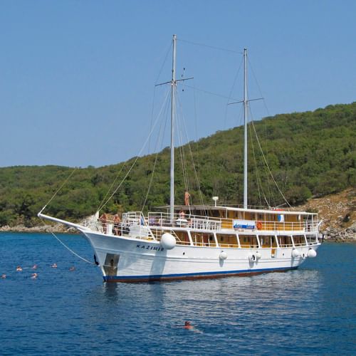 White motor sailing yacht MS Kazimir with two masts anchored in calm blue water. Green hills in background, swimmers visible in water.
