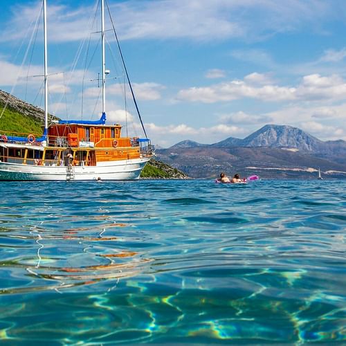 MS Mirabela, a wooden sailing ship with white masts, anchored in crystal-clear turquoise water. Mountains and green hills visible in background.